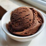 A perfectly scooped ball of rich, dark brown chocolate gelato resting in a small white bowl, illuminated by sunlight.