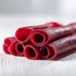 Close-up of several bright red, rolled pieces of Homemade fruit leather stacked on a white wooden surface.