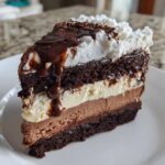 A decadent slice of 3-layer chocolate ice cream cake topped with whipped cream and fudge sauce.