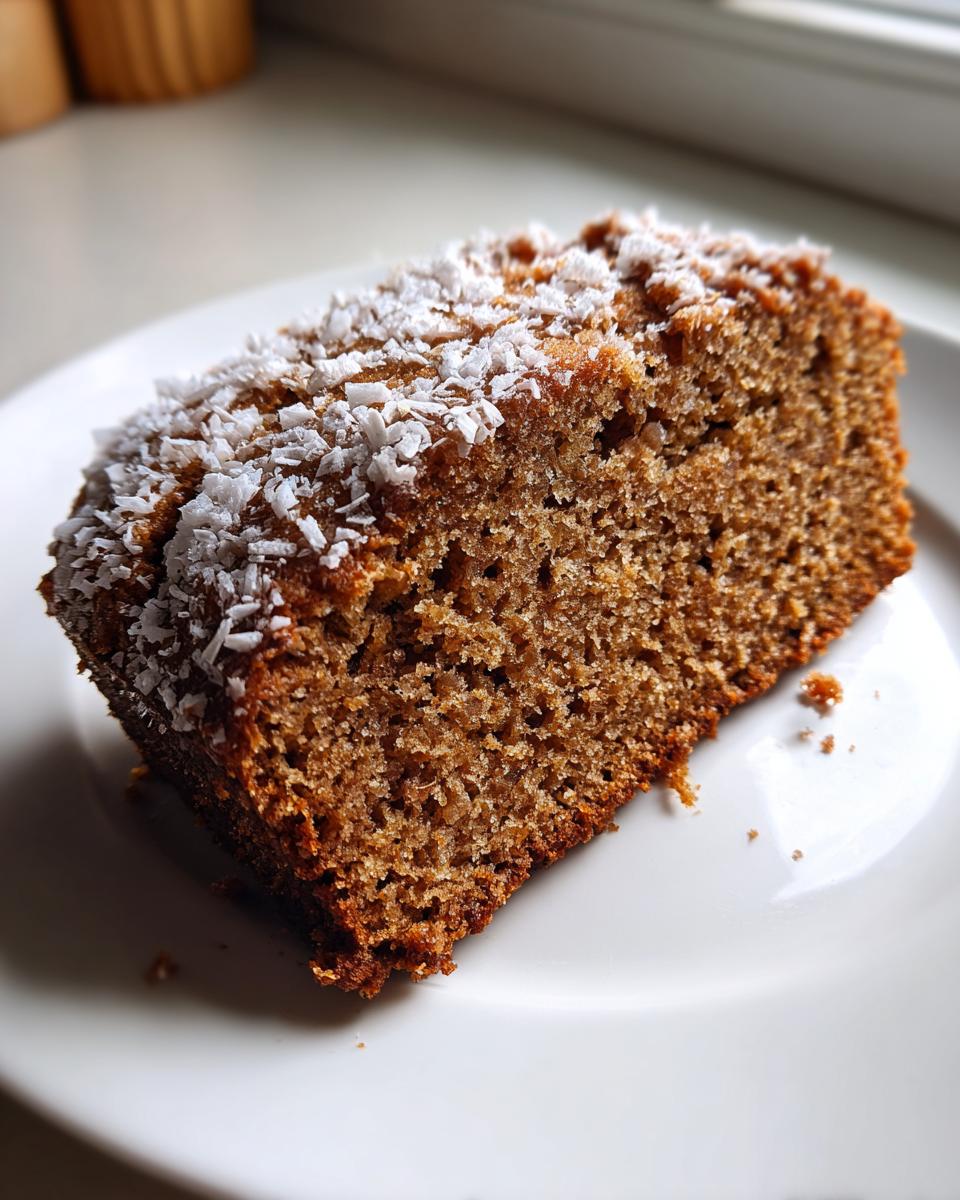 A moist slice of spiced applesauce cake topped with shredded coconut flakes, resting on a white plate.