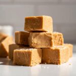 A stack of creamy, light brown peanut butter fudge squares sprinkled with coarse sea salt.