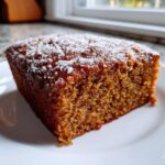 A moist slice of dark brown spiced applesauce cake dusted with coarse sugar, sitting on a white plate.