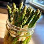 Close-up of bright green pickled asparagus spears standing upright in a glass jar with garlic cloves and red pepper flakes.