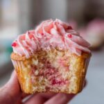 A close-up of a strawberry cupcake cut in half, showing the moist cake interior with berry swirls and pink frosting.