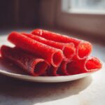 Close-up of a stack of bright red, rolled Strawberry fruit roll up snacks resting on a white plate near a window.