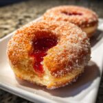 Close-up of a sugared strawberry jelly donut with a bite taken out, revealing bright red filling.