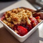 Close-up of a single serving of warm Strawberry Rhubarb Crisp in a white dish, showing juicy fruit and a buttery crumble topping.