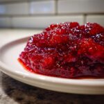 A close-up, glossy mound of vibrant red strawberry rhubarb jam served on a white plate.