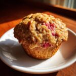 A freshly baked strawberry rhubarb muffin with a golden-brown, textured top showing bright red fruit pieces.