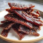 A close-up stack of glistening, dark brown Teriyaki turkey jerky pieces sprinkled with spices on a light plate.