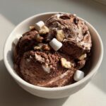 Close-up of two scoops of rich chocolate Rocky Road ice cream topped with marshmallows and chopped nuts in a white bowl.