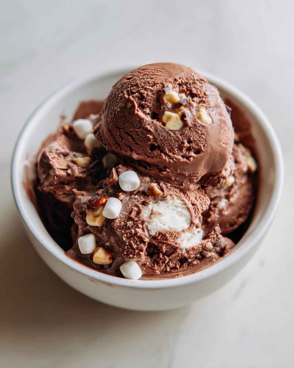 Close-up of thick scoops of chocolate Rocky Road ice cream topped with marshmallows and nuts in a white bowl.