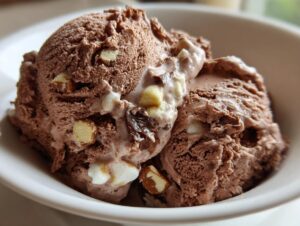 Close-up of two thick scoops of chocolate Rocky Road ice cream with visible marshmallows and nuts in a white bowl.