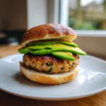 Close-up of a juicy turkey burger with sliced avocado stacked on a toasted bun, served on a white plate.