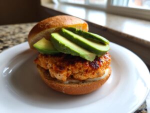 A close-up of a juicy turkey burger with sliced avocado stacked on a toasted bun, served on a white plate.