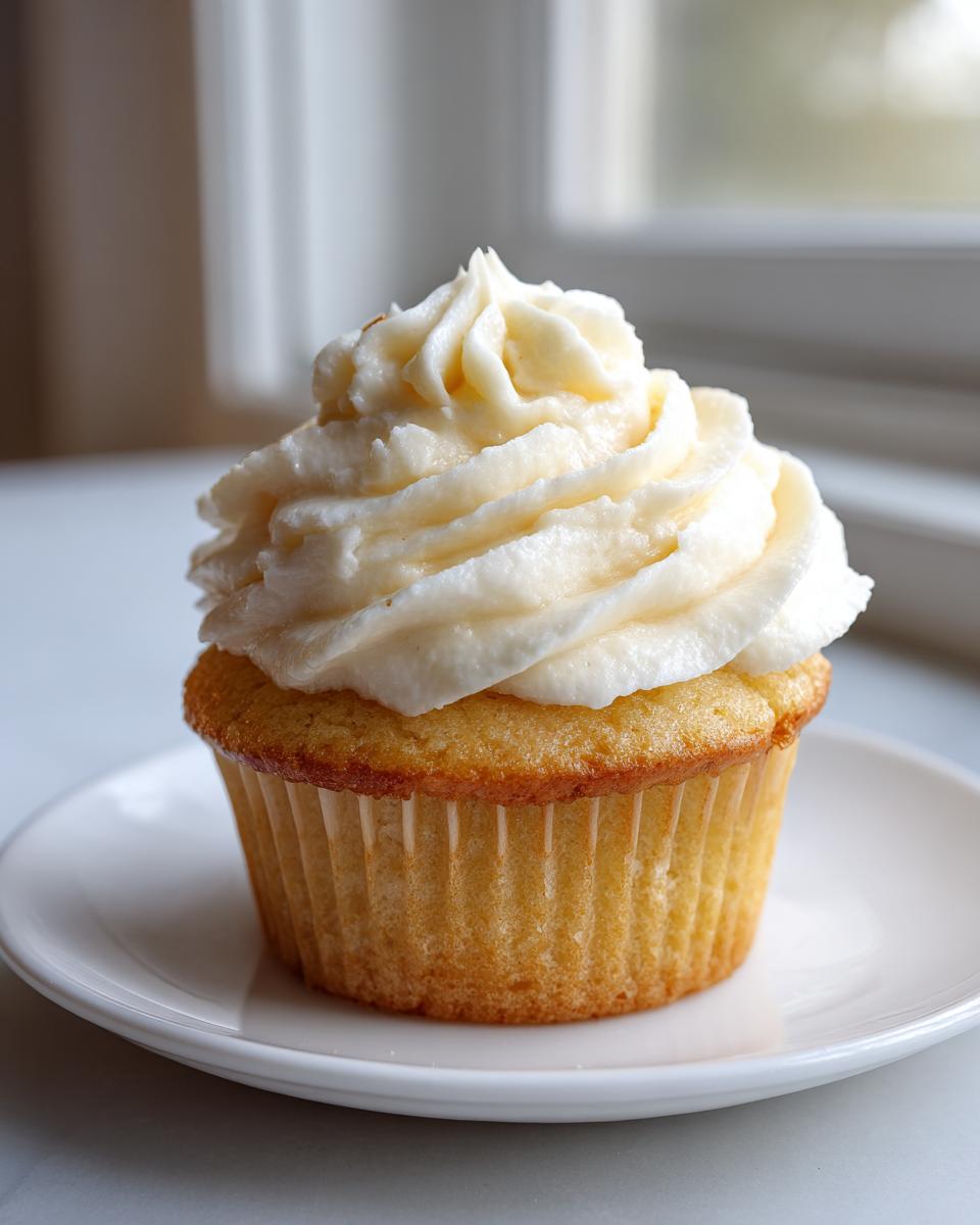 A single, perfectly baked vanilla cupcake topped high with swirled vanilla buttercream frosting, sitting on a white plate.