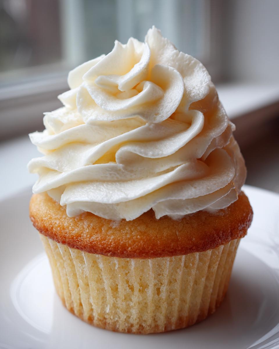 A single, perfectly baked vanilla cupcake topped with a tall swirl of creamy white buttercream frosting.