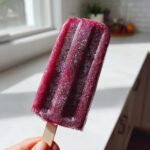 A hand holding a deeply purple, frozen grape popsicle with visible ice crystals in bright sunlight.