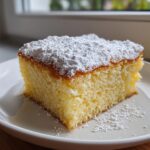 A moist slice of warm milk cake topped generously with white powdered sugar, served on a white plate.